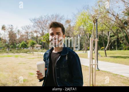Giovane uomo che indossa una giacca jean che tiene una tazza di caffè e sorride mentre si trova nel parco cittadino. Pausa caffè. Foto Stock