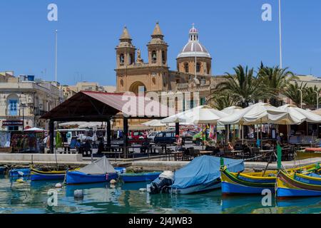 Marsaxlokk, Malta - Giugno 8th 2016: Barche da pesca maltesi tradizionali chiamate Luzzu ormeggiate nel porto con la chiesa parrocchiale sullo sfondo. Foto Stock