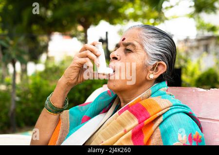 donna anziana che usa l'inalatore di asma mentre si siede al parco a causa di allergia - concetto che mostra gli effetti di inquinamento, illnes e malattia Foto Stock