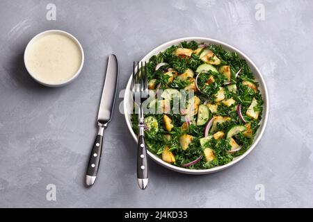 Insalata Kale con patate al forno, cetrioli, cipolle rosse e salsa di yogurt senape su sfondo grigio, vista dall'alto Foto Stock