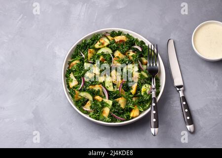 Insalata Kale con patate al forno, cetrioli, cipolle rosse e salsa di yogurt senape su sfondo grigio, vista dall'alto Foto Stock