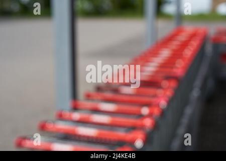 Sfocato. Una fila di carrelli per lo shopping con maniglie rosse da un negozio di alimentari (Penny). Colonne grigie e posto auto sullo sfondo. Vista laterale posteriore. Alto Foto Stock