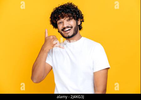 Chiamami bambino. Uomo positivo indiano o arabo millenario che tiene la mano vicino all'orecchio, mostrando il segno del telefono con le dita, chiedendo il numero, in piedi su sfondo arancione isolato, guarda la macchina fotografica, sorridente Foto Stock