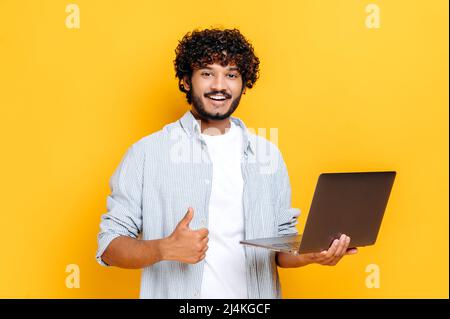 Positivo attraente ragazzo arabo o indiano con capelli ricci, indossare abiti casual, tenere un portatile aperto in mano, guardare la fotocamera, mostrando i pollici verso l'alto gesto, si leva su sfondo arancione isolato Foto Stock