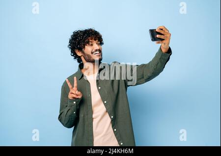 Allegro felice indiano o arabo eccitato ragazzo con gladden volto espressione, in abiti casual elegante, facendo selfie girato su smartphone e sorride nella fotocamera, si leva su isolato sfondo blu Foto Stock