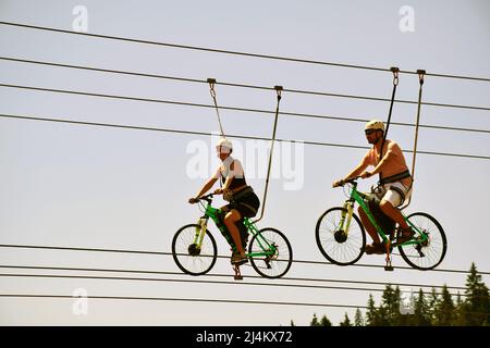 All'attrazione, un ragazzo e una ragazza cavalcano in bicicletta su corde appese e sorridono. Fotografato dal basso contro il cielo blu Foto Stock