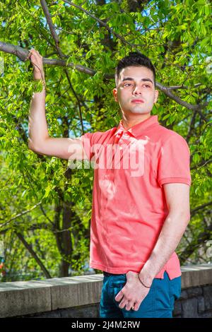 Vestendo in una polo rossa, tenendo un ramo grande di un albero, un giovane ragazzo bello sta in piedi fuori in primavera, pensando. Foto Stock