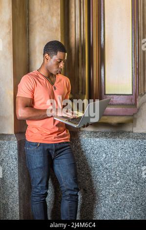 Vestendo in una camicia a V a manica corta arancione chiaro, jeans, un giovane studente nero bello è in piedi da una piccola finestra, lavorando su un computer portatile Foto Stock