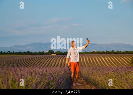 L'uomo brutale bello con capelli lunghi bruna rende selfie nel campo della lavanda in provenza vicino a Valensole al tramonto, Francia, chiaro sole Foto Stock