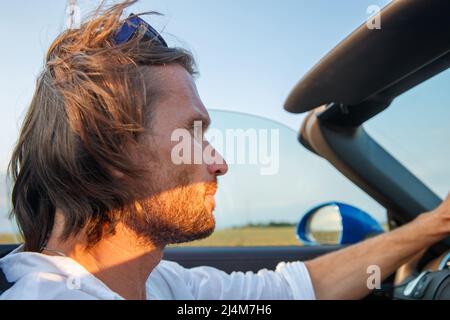 L'uomo brutale bello con capelli lunghi e una barba facile a una macchina senza top al tramonto, cabriolet, colore blu, occhiali da sole Foto Stock