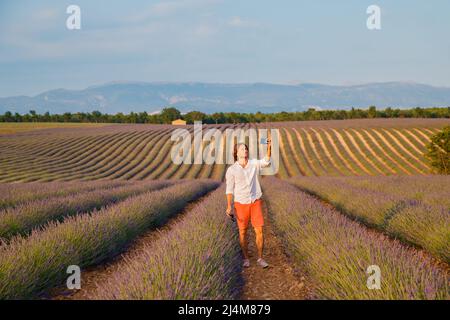 L'uomo brutale bello con capelli lunghi bruna rende selfie nel campo della lavanda in provenza vicino a Valensole al tramonto, Francia, chiaro sole Foto Stock
