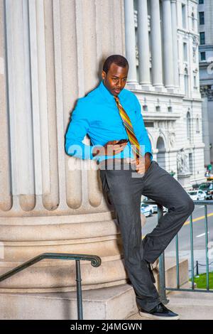 Vestendo in una camicia blu chiaro, pantaloni grigi, scarpe di pelle, una cravatta motivo, un giovane uomo d'affari nero è in piedi fuori di un edificio ufficio, controllo Foto Stock