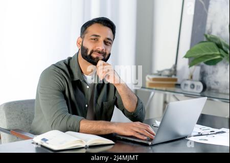 Ritratto di un carismatico uomo indiano o arabo, vestito elegantemente, imprenditore, freelancer o designer, seduto su una scrivania in un ufficio moderno, guardando la macchina fotografica, sorridente amichevole Foto Stock