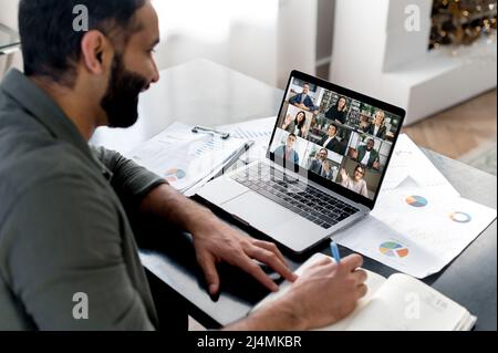 Briefing aziendale. Il ragazzo indiano che guarda lo schermo del laptop, parla in videochiamata con il gruppo aziendale multinazionale, tiene una conferenza, una riunione virtuale, un brainstorming con i colleghi online, prende appunti Foto Stock