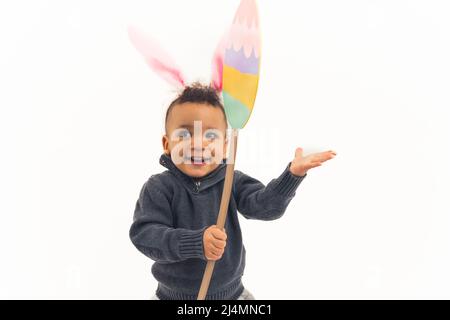 Concetto di pausa di Pasqua. Innocente curioso ragazzo biraciale ragazzo ridendo mentre indossando le orecchie di coniglietto di furry e tenendo l'uovo di Pasqua di cartone su sfondo bianco. Foto di alta qualità Foto Stock