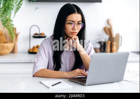 Ha messo a fuoco la ragazza asiatica graziosa con gli occhiali, la giovane donna cinese, il freelancer o lo studente, che lavora o studia a casa usando il laptop, guardando intentamente lo schermo del laptop, studiando le informazioni Foto Stock