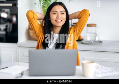 Felice ragazza cinese, freelancer, lavorando da casa, si siede in cucina sul posto di lavoro, prende una pausa dal lavoro, mette le mani dietro la testa, guarda al lato, sogni di riposo, pensa, sorride Foto Stock