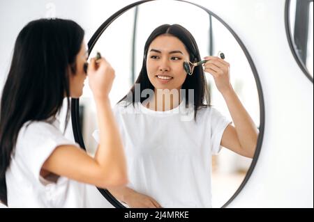 Felice asian brunette bella ragazza in t-shirt, si alza di fronte a uno specchio a casa, fa la sua routine di bellezza mattina, si prende cura della sua pelle facciale, utilizza un rullo di pietra facciale, previene le rughe, sorriso Foto Stock