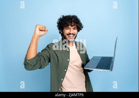 Allegro ragazzo arabo o indiano felice in abiti casual, studente o freelance, tiene un laptop aperto in mano, si rallegra nella vittoria, gesturing con il suo pugno, in piedi su un isolato sfondo blu Foto Stock