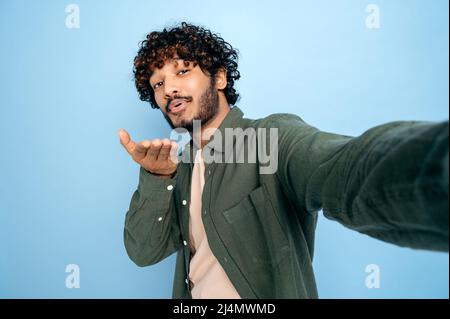 Felice positivo indiano o arabo ragazzo eccitato con, in abbigliamento casual, facendo selfie girato sul cellulare e sorrisi nella fotocamera, fa un bacio d'aria, si leva su isolato sfondo blu Foto Stock