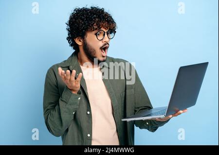 Sorpreso indovinato astuto indiano o arabo ragazzo con occhiali, in abiti casual, tiene un laptop aperto, sembra confuso allo schermo, gesturing mano, si leva su isolato sfondo blu Foto Stock
