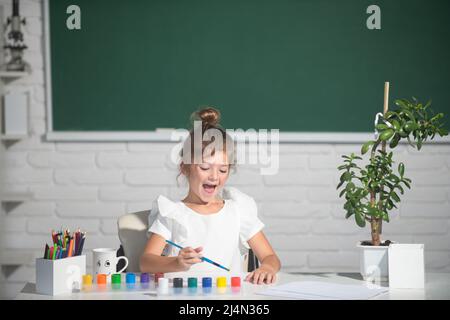 Bambina attira in aula seduto a un tavolo, divertendosi sulla lavagna di fondo della scuola. Istruzione creativa per i bambini. Foto Stock