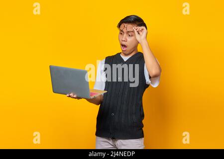 Scioccato asiatico giovane uomo utilizzando il portatile e prendendo di occhiali isolati su sfondo giallo Foto Stock
