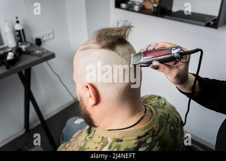 Un giovane uomo in uniforme militare rasa la testa calva per il servizio militare. Un ragazzo con una barba ottiene un taglio di capelli ad un negozio di barbiere. Foto Stock