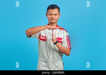 Ritratto del giovane meccanico asiatico che mostra il gesto di timeout su sfondo blu Foto Stock