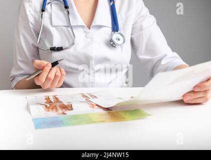 Dottore che tiene carte e studia anatomia. Educazione medica e concetto di conduzione della ricerca. Donna in camice da laboratorio con stetoscopio seduto a tavola e documenti di analisi. Foto di alta qualità Foto Stock