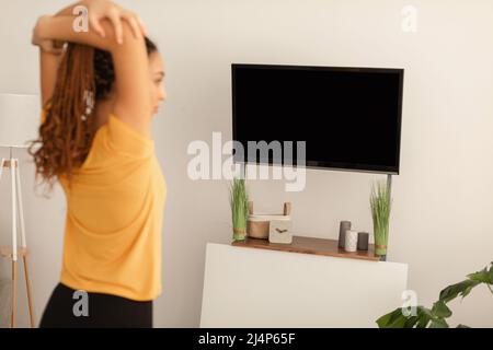 Black Woman Stretching Arms esercitandosi guardando schermo della televisione all'interno Foto Stock