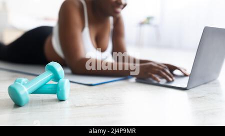 Plump giovane donna nera sdraiata sul tappeto yoga e guardare video online di sport sul portatile a casa, concentrarsi su manubri Foto Stock