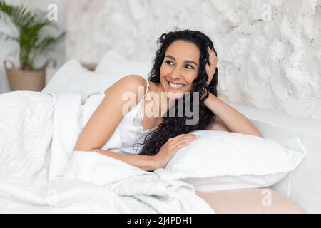 Soddisfatto giovane donna bruna caucasica con capelli ricci lunghi in pigiama gode di tempo libero, sdraiato a letto Foto Stock