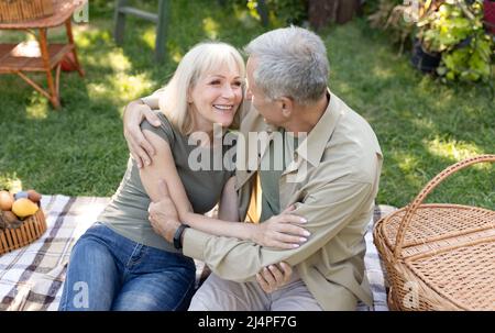 I coniugi più grandi sono entusiasti di fare un picnic in giardino, di abbracciare e ridere, seduti insieme su coperta all'aperto Foto Stock