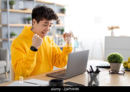 L'uomo asiatico eccitato che usa il pc festeggia il successo scuotendo i pugni Foto Stock