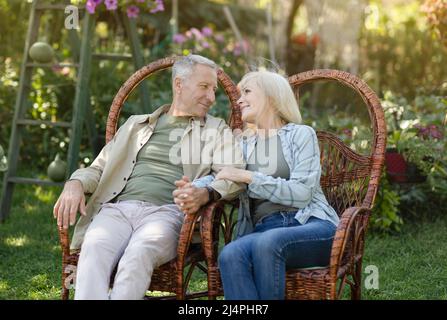Coppia sposata felice senior che riposa all'aperto in giardino, seduto su sedie di vimini e guardandosi l'un l'altro Foto Stock