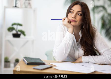 Una ragazza creativa entusiasta dai capelli scuri in una elegante camicia bianca si siede su una scrivania e attinge nel suo notebook o prende appunti nel suo diario, nel suo co Foto Stock