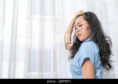 Una donna di occhiali asiatici in camicia blu pone da affettare i suoi capelli indietro di fronte alla tenda alleggerente. Foto Stock