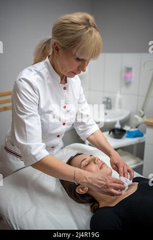 Medico Beutician strofinando il viso del paziente con tamponi di cotone. Ritratto di donna sulla pulizia del viso procedura da cosmetologo donna in clinica di bellezza. Bellezza Foto Stock