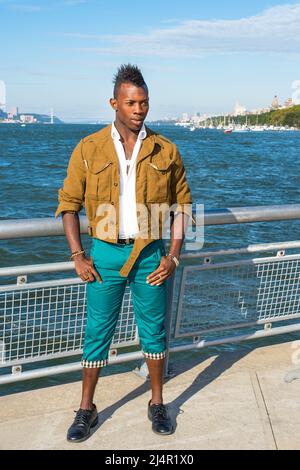 Vestendo in una giacca di lana gialla, pantaloni verdi e scarpe di pelle nera, un giovane ragazzo nero con capelli mohawk è in piedi da un fiume e con fiducia l Foto Stock