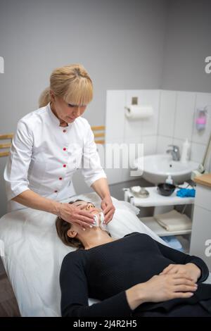 Medico Beutician strofinando il viso del paziente con tamponi di cotone. Ritratto di donna sulla pulizia del viso procedura da cosmetologo donna in clinica di bellezza. Bellezza Foto Stock