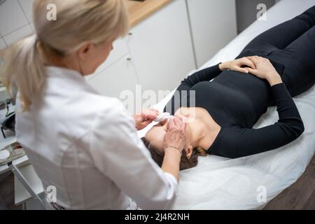 Medico Beutician strofinando il viso del paziente con tamponi di cotone. Ritratto di donna sulla pulizia del viso procedura da cosmetologo donna in clinica di bellezza. Bellezza Foto Stock