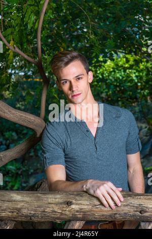 Vestendo in una maniche lunghe grigie con la camicia Henley roll-tab, un giovane ragazzo bello è in piedi in una zona di legno e guardando attentamente voi. Foto Stock