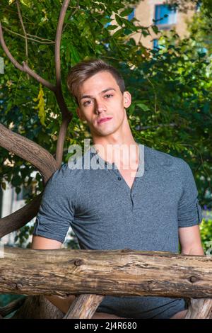 Vestendo in una maniche lunghe grigie con la camicia Henley roll-tab, un giovane ragazzo bello è in piedi in una zona di legno e ti guarda con fascino. Foto Stock