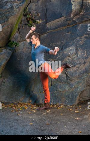 Vestendo in maniche lunghe grigie con camicia Henley con linguetta di rotolamento, jeans rossi e scarpe da stivale in pelle marrone, un ragazzo giovane sta allungando le braccia e le gambe, exercisi Foto Stock