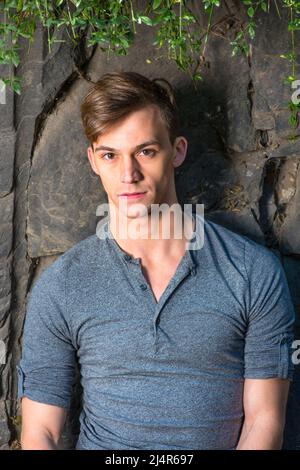 Vestendo in una maniche lunghe grigie con rotolo-linguetta camicia Henley, un ragazzo bello giovane è in piedi da rocce, foglie verdi sulla parte superiore, e affascinante lookin Foto Stock