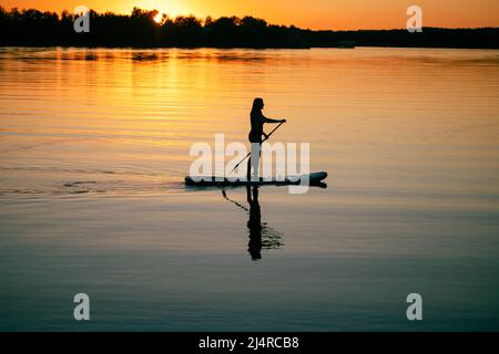 Femmina sup boarder con rospo in mano che si muove intorno sul fiume con meraviglioso tramonto sullo sfondo e riflessione sulla superficie d'acqua in estate. Attivo Foto Stock