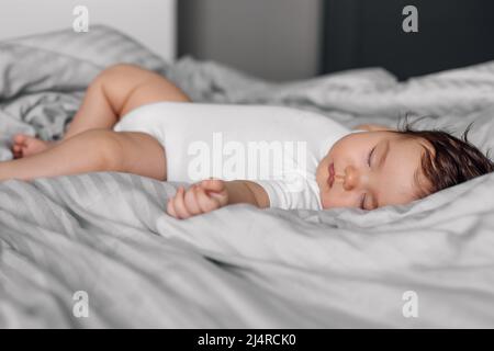 Ritratto del bambino nel bodysuit di sonno che giace sulla coperta sul letto. Dormire bambino in camera da letto, vedere dolce sogno. Sfondo grigio, messa a fuoco morbida, copia gratuita Foto Stock