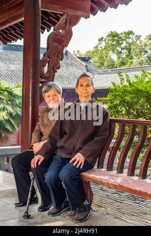 Uomo anziano e donna seduti su una lunga panca per fare una pausa. Una coppia anziana, di 80 anni, è seduta su una panchina all'interno di un tempio buddista mentre si trova di fronte Foto Stock