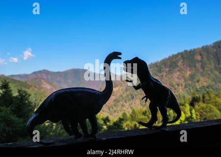 Due dinosauri, un brontosauro e un t rex, si confrontano tra loro in silhouette con montagne fuori fuoco in lontananza Foto Stock